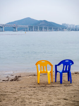 View of a yellow and a blue plastic chair placed on the sandy beach with the Gwangan Bridge visible in the background in Busan, Busan, South Korea.