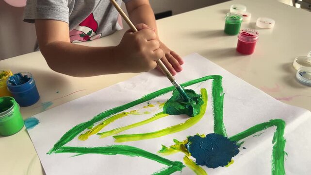 Child paints a large green tree with a brush on paper, creative preschool art activity and early learning at home.