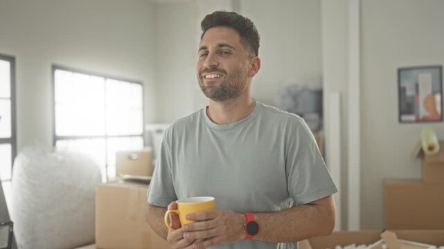 Man holding yellow mug, hands visible, smiling while standing among moving boxes in a building; contentment transition.