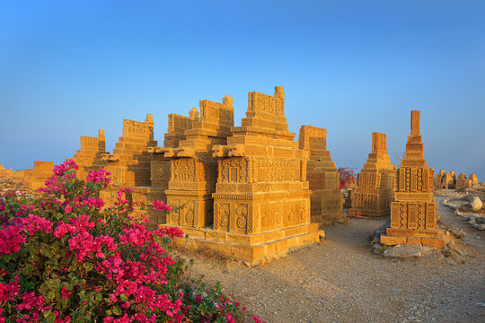 Karachi, Pakistan - 22 January 2010: View of the historic Chaukhandi Tombs featuring intricately carved sandstone structures and vibrant pink bougainvillea flowers under a clear blue sky.