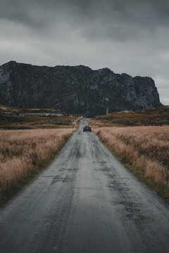 View of a car driving along a gravel road towards the rugged mountains of Nesland, surrounded by dry grass under a dramatic sky in Flakstad Municipality, Nordland, Norway.
