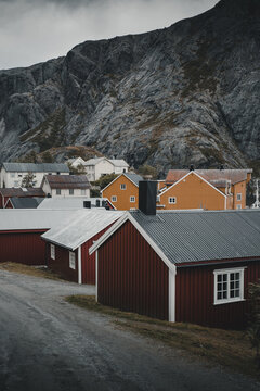 Nusfjord, Norway - 07 September 2022: View of Nusfjord featuring traditional red and yellow wooden rorbu cabins nestled against a dramatic rocky mountain backdrop under a cloudy sky.