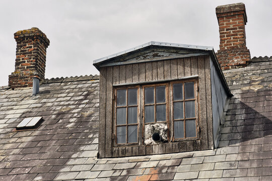 Viborg, Denmark - 07 August 2026: View of an old roof featuring a wooden dormer window, weathered slate tiles, and two brick chimneys against a cloudy sky.