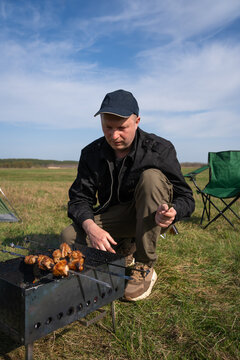 Man squatting outdoors in a green field, cooking meat on metal skewers over a portable charcoal barbecue while enjoying leisure time under a clear blue sky. Vertical photo