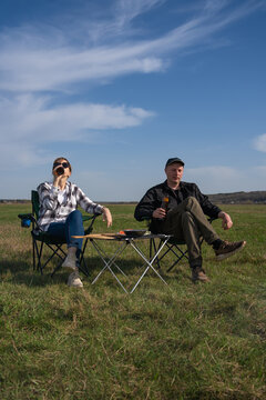 Couple enjoying a relaxing picnic moment, sitting in camping chairs beside a tent, drinking beer together in a vast green field under a clear blue sky. Vertical photo
