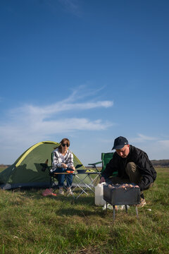 Couple preparing food and grilling barbecue meat on a portable grill while camping in a green field under a clear blue sky, experiencing outdoor leisure activities. Vertical photo