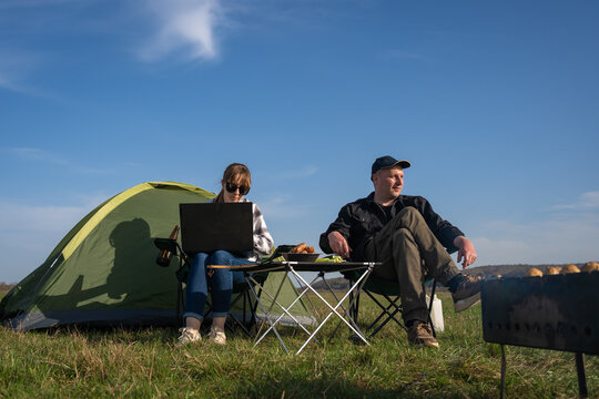 Couple camping in a sunny meadow: woman focused on laptop working remotely while man relaxes by the tent, enjoying nature, freedom and a peaceful outdoor office