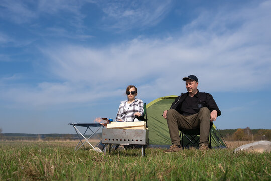 Couple resting and barbecuing near a green tent in a wide open field under a blue sky, enjoying outdoor leisure activities and a natural landscape during their summer vacation