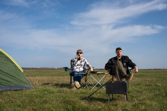 Young couple camping in a sunny meadow, sitting by a tent and portable grill, relaxing together under blue skies while enjoying outdoor cooking, bonding and fresh air