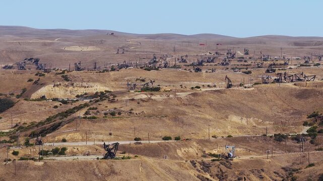 Aerial view of multiple pump jacks extracting crude oil across dry arid hills in a California oil field. The machinery pumps steadily along dusty access roads. Oil tariffs, trade war, price plunge.