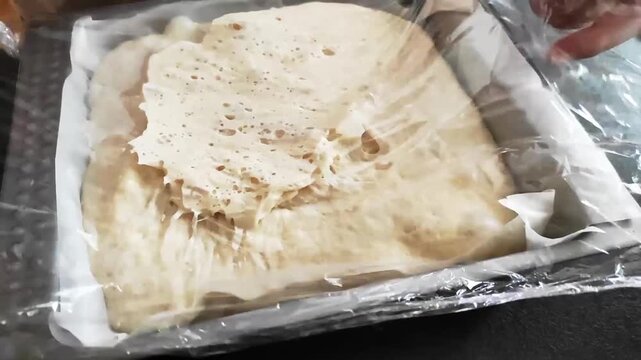 Close-up of sticky, bubbly focaccia dough being uncovered by peeling off plastic wrap in a parchment-lined baking tray before baking.