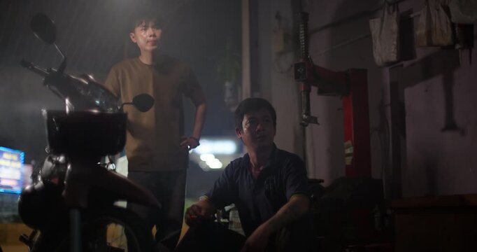 Two mechanics gather beside a moped while watching a football game at the repair shop at night, eager anticipation and cheering with happiness to celebrate
