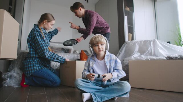 The couple is packing things while their son sits on the floor playing a video game. Cardboard boxes are around them.
