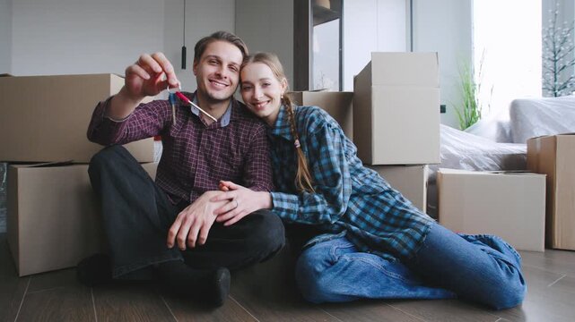 A couple sits on the floor surrounded by cardboard boxes in their new home, smile while holding a set of keys.