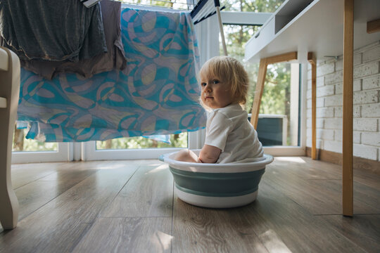 Cute toddler sitting inside a plastic laundry basin on the floor under drying clothes at home. Childhood moment showing a little child playing with everyday household objects while laundry dries