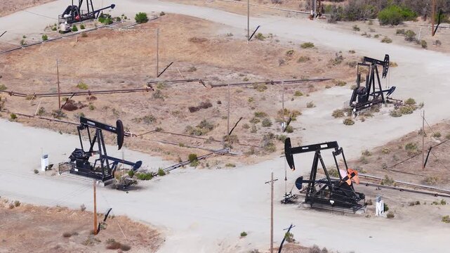 Aerial view shows pump jacks steadily extracting crude petroleum within an arid oil field, featuring pipelines and dirt service roads connecting the industrial wells across the dry terrain.