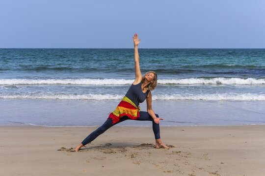 A woman doing yoga on a sandy beach by the sea. Parshvakonasana or Side Angle. Treen Cove, Cornwall, England, Great Britain
