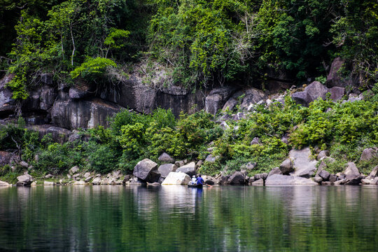 Meghalaya, India - 31 March 2023: View of a small boat with two people rowing on a calm river surrounded by lush green cliffs and large boulders.