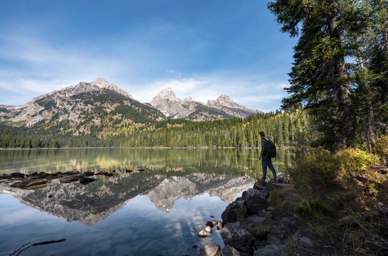 Young man standing by a lake, reflection in Taggart Lake, view of Teton Range mountain, peaks Grand Teton and Teewinot Mountain, Grand Teton National Park, Wyoming, USA