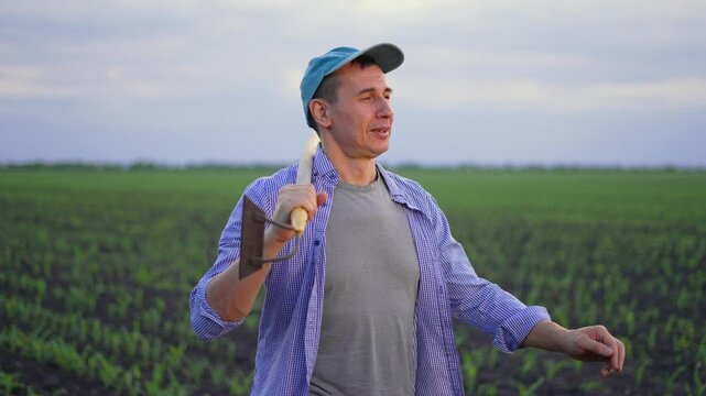 Cheerful farmer stands in green corn field holding hoe on shoulder. Farmer in cap and plaid shirt talks and gestures with hand. Agriculture worker rests in cultivated field of young crop at dusk.