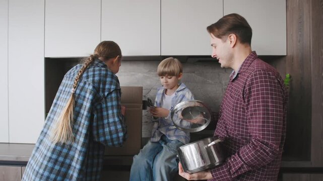 A family stands in the kitchen surrounded by cardboard boxes, playing with kitchenware and utensils. They are having fun unpacking their belongings in their new apartment.
