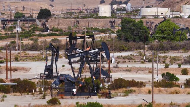 Aerial view tracks active pumpjacks pumping petroleum across an arid California oil field, surrounded by storage tanks, utility poles, and dry terrain. Oil tariffs, trade war, price plunge.