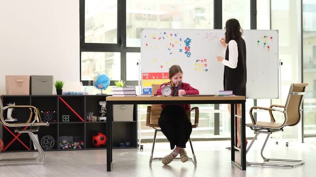 Experienced English teacher writes lesson topic on large whiteboard. Diligent student sits at desk and takes detailed notes in open exercise book