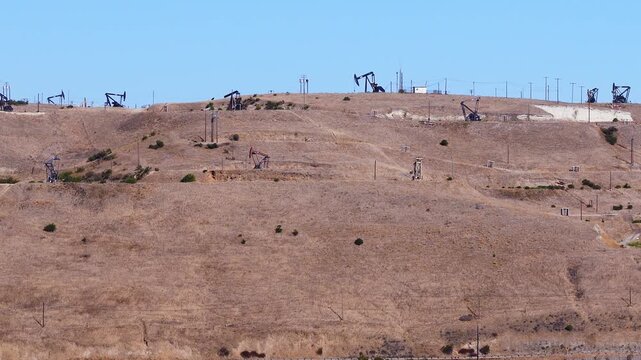Aerial view showing multiple working pump jacks extracting petroleum across dry arid hillsides in a California oil field during broad clear daylight conditions.