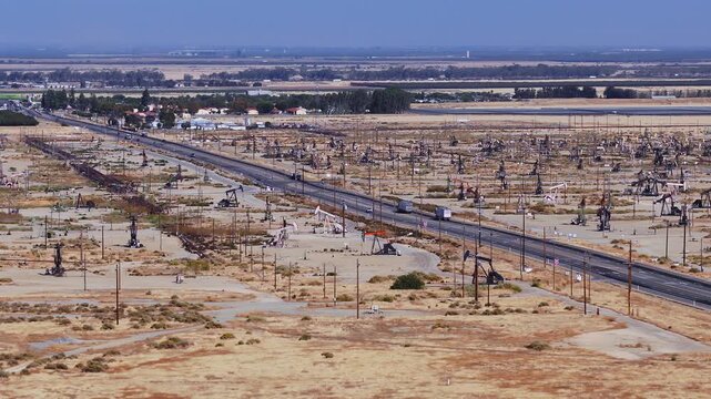Aerial view shows numerous oil pump jacks extracting petroleum beside a highway in a California desert field, with trucks and cars traveling along the asphalt road on a clear sunny day.