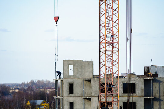 Construction site background. A tall orange tower crane beside an unfinished concrete building. An unidentified worker on the edge and bare trees in the distant landscape, view from height