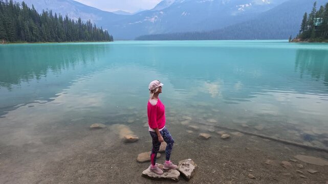 top view of woman stands on shoreline of Emerald Lake in Yoho National Park, gazing over turquoise water and evergreen forests with mountain peaks, serene travel moment in British Columbia, Canada