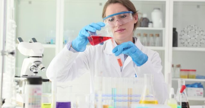 Woman researcher pours red liquid from beaker into test tube in lab slow motion. Female chemist conducts analysis of fluid in scientific facility