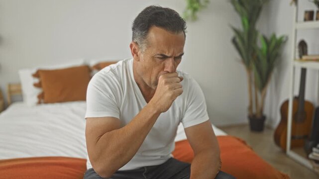 Man clutching chest and covering mouth while seated on bed in bedroom with plant, guitar and orange pillows visible; pain concern.