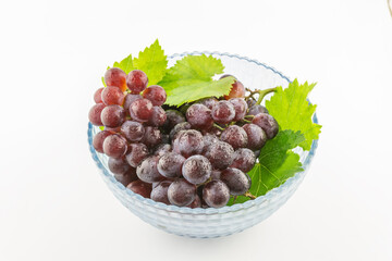 Fresh red and purple grapes with water droplets, accompanied by green vine leaves, placed in a clear glass bowl against a white background. © tai