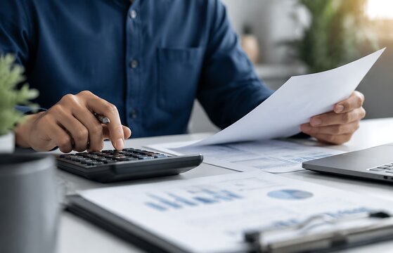 Person calculating financial documents at a desk in an office with man blue shirt uses calculator while reviewing business charts paperwork desk, business professional