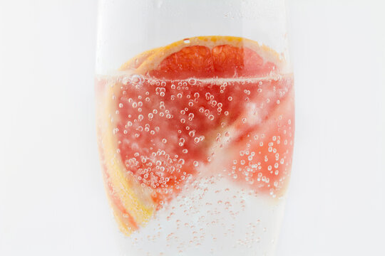 Close-up of fresh grapefruit slices submerged in bubbly sparkling water in a clear glass, against a plain white background.