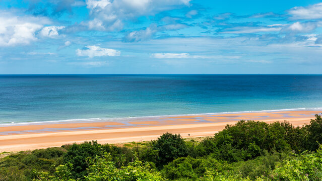Colleville-sur-Mer, Normandie, France - 18 June 2026: View of Omaha Beach with its sandy shoreline, calm blue ocean, and lush green vegetation under a bright sky with clouds.