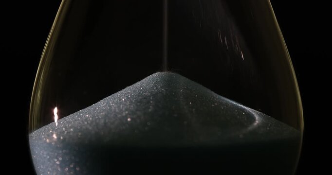 Shimmering white sand poured down within hourglass in dark space. Vintage tool with glitter powder measures time periods. Lifecycle concept
