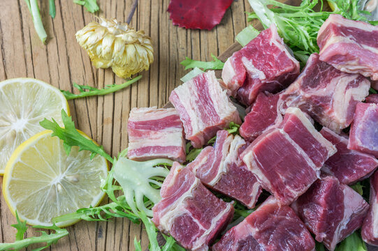 Raw marbled beef chunks arranged with lemon slices and fresh greens on a wooden surface, food preparation scene.