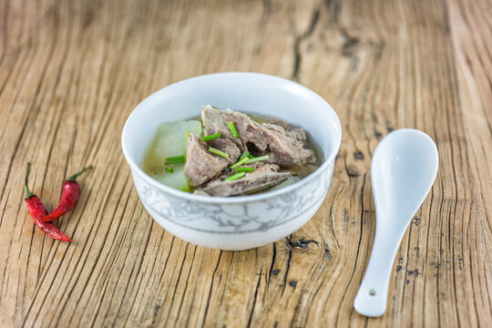 A bowl of savory stewed meat and winter melon soup with chopped scallions, served with a spoon and red chilies on a rustic wooden table.