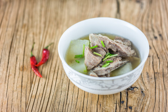 A bowl of pork and winter melon soup garnished with scallions, placed on a wooden table with two red chilies beside it.
