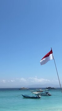 A clear blue sky over a tranquil sea with small boats anchored near the shore. An Indonesian flag waves gently in the breeze.