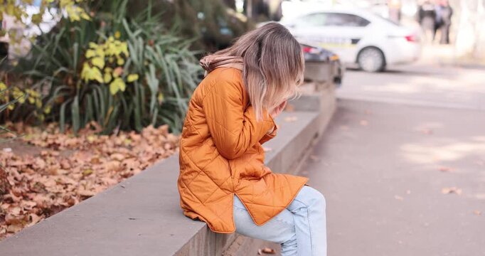 Woman sneezes while sitting on park stone parapet near autumn street. Female person blows nose with tissue covering face during sudden cold symptoms