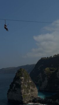 Person zip-lining over a scenic landscape with cliffs and a blue ocean in the background. Clear sky and lush greenery are visible.