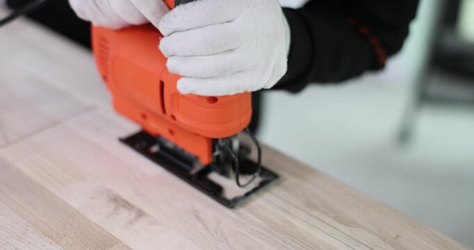 Gloved hands guide orange electric jigsaw across wooden board. Powered blade cuts straight path while tool moves steadily through timber surface
