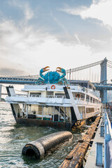 Fototapeta premium Crab sculpture above a moored vessel and floating fender. A white boat sits beside a pier in New York City, with a black fender in the water and a bridge behind.