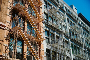 Exterior metal staircases across rows of building windows. Orange and gray escape stairs cast shadows across masonry facades in New York City under direct sunlight. © Vadim