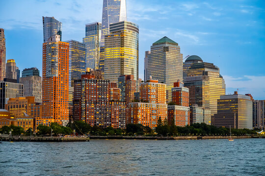 Waterfront apartments below glass towers with illuminated windows. Brick residential blocks line the river in New York City, with lit office floors and geometric roofs above.