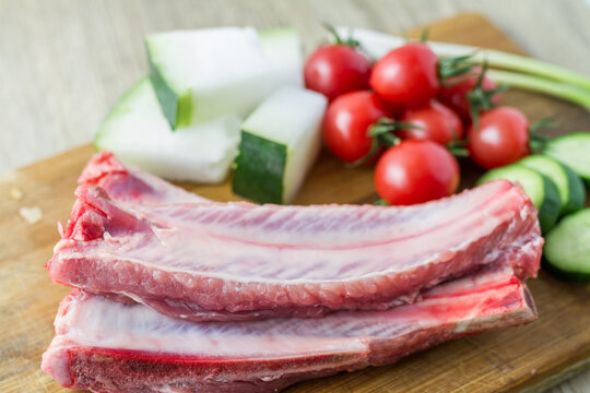 Raw pork ribs and assorted fresh vegetables placed on a wooden cutting board for cooking preparation.