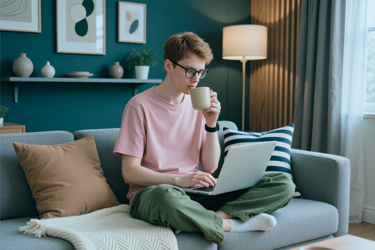 Young adult Caucasian man sitting on sofa, typing on laptop and drinking coffee. Image represents remote work, online education, and digital lifestyle, suitable for home office marketing